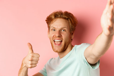 Excited redhead man tourist taking selfie and showing thumbs-up, holding camera with stretched out...