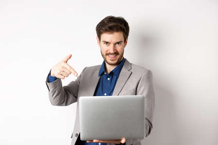 Confident young male entrepreneur working online with laptop, pointing at computer and smiling, standing on white backgroundの写真素材