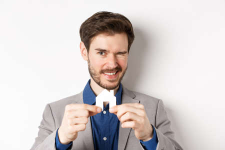 Real estate and insurance concept. Handsome man in suit winking and smiling, showing small paper house cutout, standing on white backgroundの写真素材