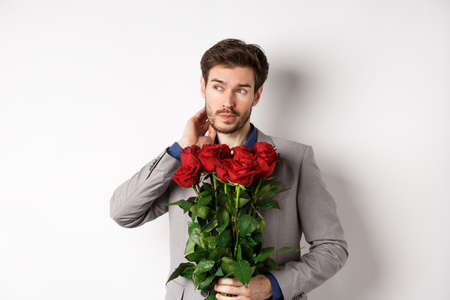 Pensive young man in suit holding bouquet of flowers, waiting for date on Valentines day, standing over white backgroundの写真素材