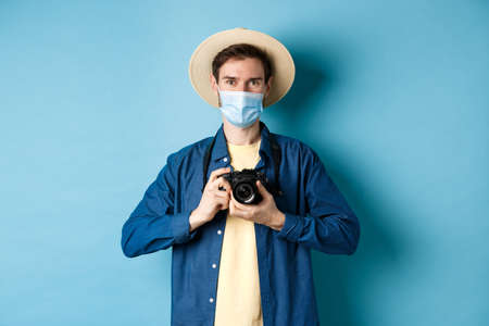 Covid-19, pandemic and travel concept. Happy guy on vacation taking pictures with camera, wearing medical mask and summer hat, blue backgroundの写真素材