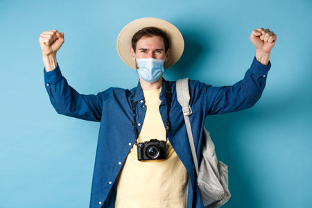 Covid-19 and summer holidays concept. Happy smiling tourist celebrating vacation, wearing face mask,...