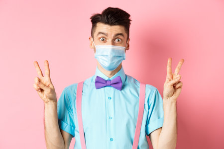 Coronavirus, healthcare and quarantine concept. Funny guy in bow-tie and face mask showing peace, victory signs and looking happy at camera, standing on pink backgroundの写真素材