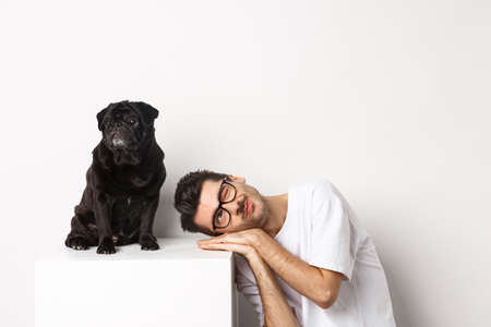 Handsome young man lay head near cute black pug, smiling and looking up at copy space, white backgroundの写真素材