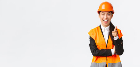 Smiling professional asian female construction manager in safety helmet and reflective jacket inviting check-out new building, wink and pointing finger at camera, showing client best estateの写真素材