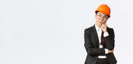 Thoughtful creative female asian chief architect, construction engineer thinking, wearing safety helmet and suit, pondering best choice for building, standing white backgroundの写真素材