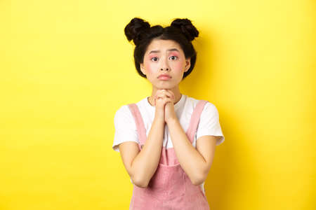 Hopeful asian stylish girl holding hands in begging sign, waiting with hope, asking for favour or help, standing on yellow backgroundの写真素材