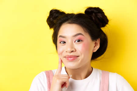 Beauty. Stylish asian girl with bright glamour makeup, touching soft and shiny facial skin, smiling happy at camera, standing on yellow backgroundの写真素材