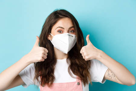 Covid-19, social distancing and lockdown concept. Young woman supports preventive measures, wearing mask respirator from coronavirus, showing thumbs up in approval, blue backgroundの写真素材