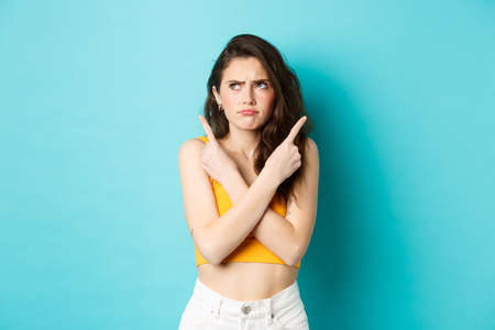 Young confused woman frowning perplexed, pointing fingers sideways and frowning displeased, need to choose, standing over blue backgroundの写真素材