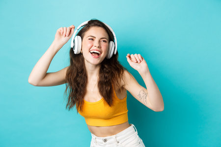 Young woman in summer clothes listening music, wearing headphones and singing along favorite song, dancing in earphones, standing against blue backgroundの写真素材