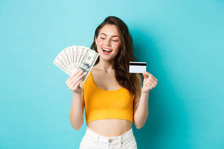 Good-looking smiling girl holding credit card, looking pleased and determined at dollar bills money, standing against blue backgroundの写真素材