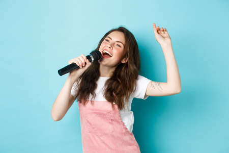 Happy young woman perform song, singer holding microphone, dancing and singing at karaoke, standing over blue backgroundの写真素材