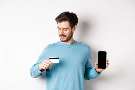 Handsome young man with beard, showing empty smartphone screen and looking at plastic credit card, standing over white backgroundの写真素材