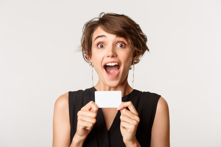 Excited woman open mouth and looking at camera, showing credit card, standing over white backgroundの写真素材