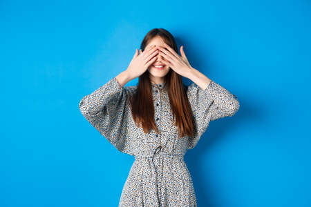 Happy smiling woman in dress cover eyes with hands, waiting for surprise, standing on blue backgroundの写真素材