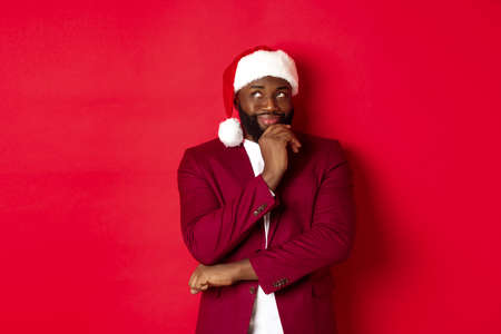 Christmas, party and holidays concept. Smiling african american man making new year plans, looking thoughtful at upper left corner, wearing santa hat, red backgroundの写真素材