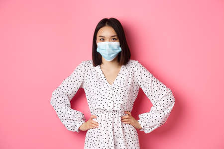 Covid-19, quarantine and lifestyle concept. Cute asian woman wearing medical mask and dress, protect herself from coronavirus, standing over pink backgroundの写真素材