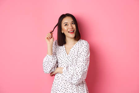 Dreamy cute asian girl thinking, imaging shopping and smiling, looking upper left corner with thoughtful smile, standing in dress over pink backgroundの写真素材