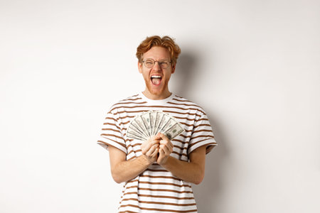 Lucky young man with red hair showing dollars, winning money and screaming of happiness, holding...