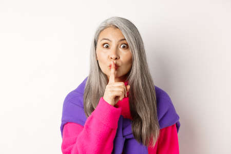 Close-up of stylish senior asian woman in hipster outfit telling hush, shushing at looking at camera, show taboo gesture, standing over white backgroundの写真素材