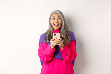 Surprised asian woman smiling at camera after reading promotion on smartphone, standing with mobile phone over white backgroundの写真素材