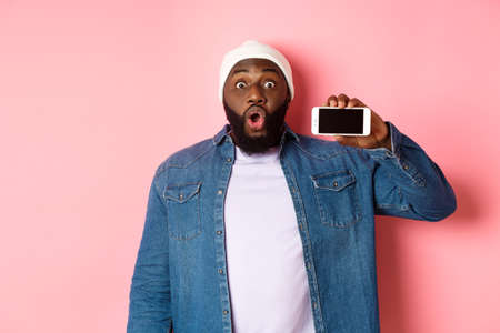 Online shopping and technology concept. Amazed Black man showing phone screen horizontally and staring at camera excited, standing over pink backgroundの写真素材