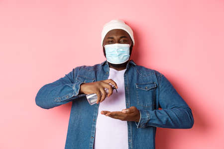Covid-19, lifestyle and lockdown concept. Smiling african-american man in face mask cleaning hands with sanitizer, using antiseptic and looking at camera, pink backgroundの写真素材