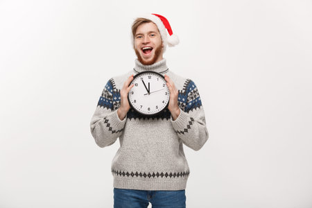 Holiday concept - Young happy handsome beard man in sweater holding white clock over white studio...