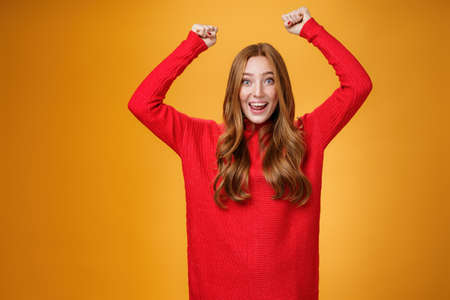 Optimistic happy and supportive ginger girl yelling cheering words, raising hands joyfully and smiling broadly triumphing, celebrating success and win, posing satisfied and excited against orange wallの写真素材