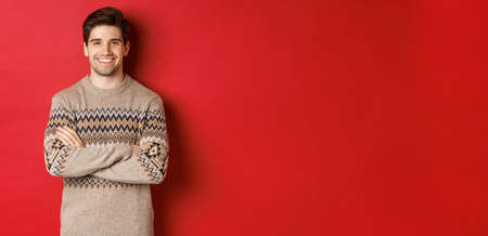 Image of handsome happy guy in christmas sweater, smiling and looking at camera, celebrating xmas holidays, standing over red backgroundの写真素材
