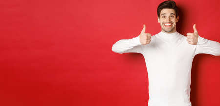 Cheerful good-looking male model in white sweater, showing thumbs-up in approval, like something good, standing over red background and smiling pleasedの写真素材