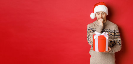 Image of worried guy in santa hat and sweater, looking indecisive at christmas gift, standing against red backgroundの写真素材