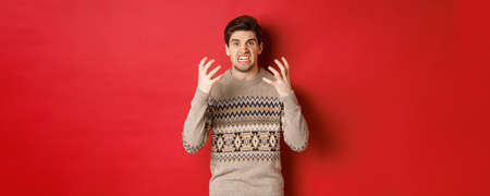 Image of angry and frustrated man in winter sweater, shouting and looking with hatred, standing upset over red backgroundの写真素材