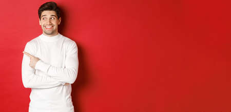Concept of winter holidays, christmas and lifestyle. Excited handsome man in white sweater, smiling, looking and pointing finger left at promo offer, standing over red backgroundの写真素材