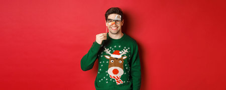 Funny man in christmas sweater and party mask, celebrating winter holidays, smiling happy, standing...