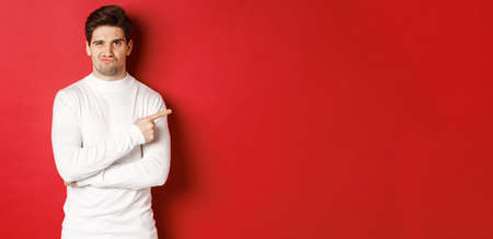 Doubtful and reluctant handsome man grimacing, pointing fingers right at something bad, standing in white sweater over red backgroundの写真素材