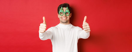 Close-up of attractive happy man in party glasses and white sweater, showing thumbs-up in approval...