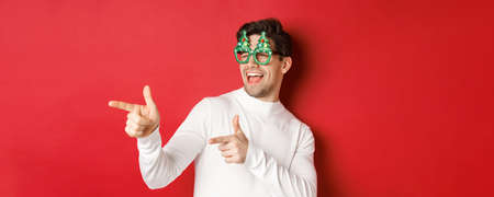 Close-up of cheeky handsome man in party glasses, pointing finger left and greeting person, wishing merry christmas, standing over red backgroundの写真素材