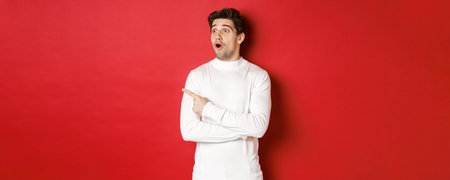 Concept of winter holidays. Portrait of impressed handsome man in white sweater, pointing and...