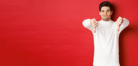 Portrait of disappointed skeptical guy, wearing white sweater, showing thumbs-down to disagree,...