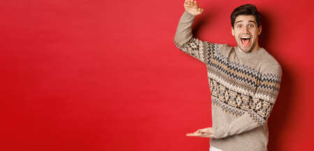 Portrait of excited and happy handsome man in christmas sweater, showing something big, holding large gift for holidays, standing over red backgroundの写真素材