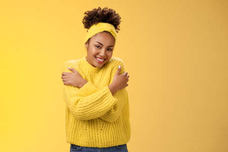 Attractive tender gentle african-american teenage girl in sweater headband hugging herself hands touch shoulders feel warm safe cozy smiling happily embracing cuddling lovely giggleの写真素材