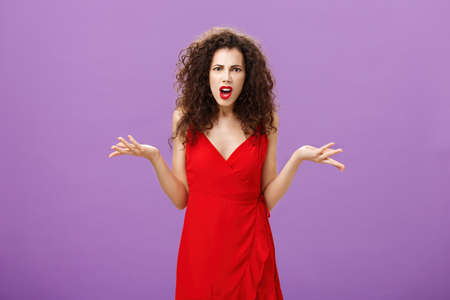 Woman being shocked guy breaking up with her during party. Concerned pissed and outraged good-looking lady in red dress with evening makeup shrugging with spread palms in clueless and questioned poseの写真素材