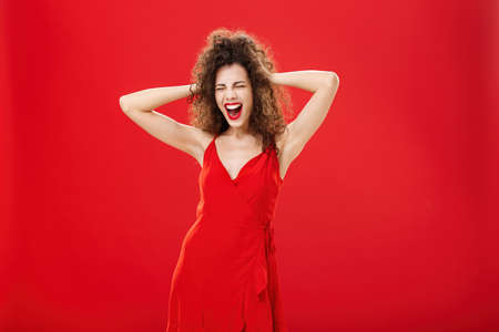 Girl just wanna have fun. Crazy adult caucasian woman with curly hairstyle in elegant dress holding hands on head yelling out loud with closed eyes and intense expression over red backgroundの写真素材
