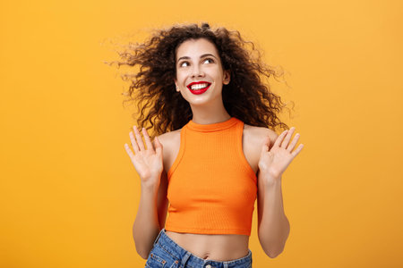 Carefree and happy stylish urban female with nose ring and curly hairstyle raising palms in clueless and indifferent gesture looking up in sun joyfully with delighted cute smile over orange backgroundの写真素材