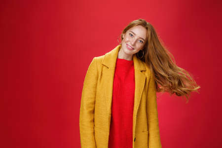 Charming redhead female with freckles waving hair smiling broadly tilting head as haircut flying in air posing delighted and carefree over red background in warm yellow coat and knitted dressの写真素材