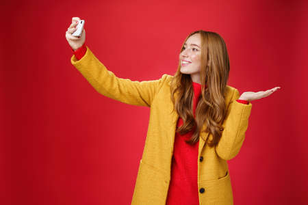 Cute redhead woman walking in park deciding take selfie near beatiful scenery or cute animal extending hand with smartphone smiling broadly at device as taking photo and pointing at somethingの写真素材