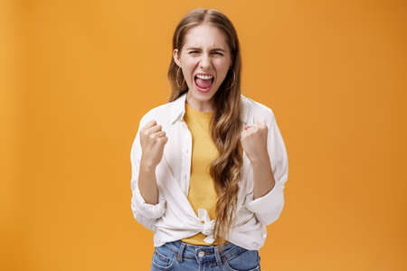 Girl feeling power overcome any trouble yelling to cheer up and boost confidence clenching raised fists in cheerful and victory gesture screaming out loud, releasing stress against orange backgroundの写真素材