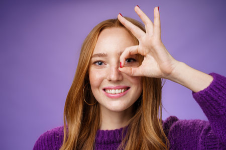 Headshot of optimistic and playful kind cute redhead female with freckles and white perfect smile showing okay or zero sign on eye, grinning as peeking through hole at camera over purple backgroundの写真素材